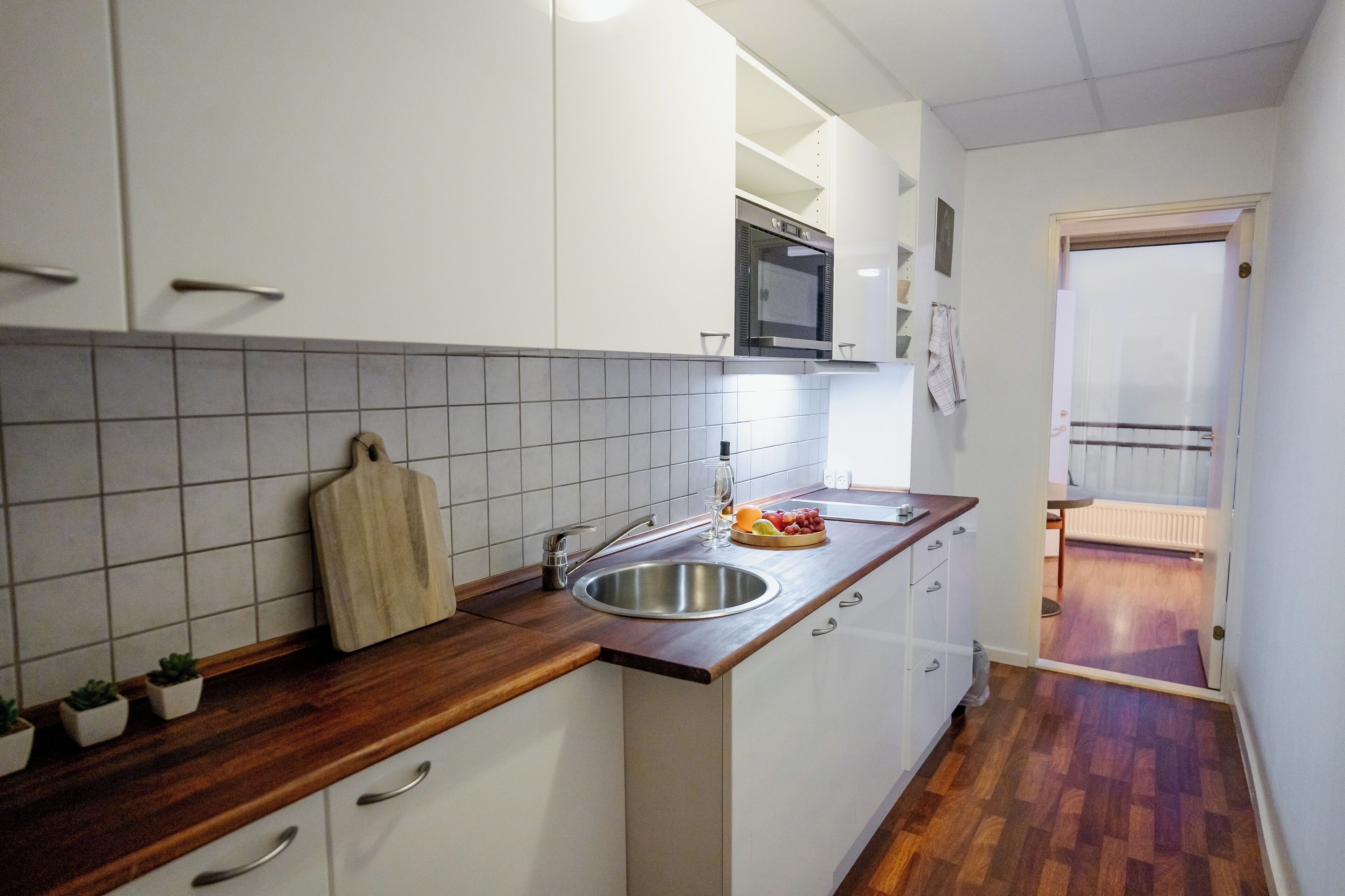 Cocina con armarios blancos, encimera de madera, pileta de acero inoxidable y un microondas encima. Hay pequeñas macetas con plantas sobre la encimera y una toalla colgada en la pared.