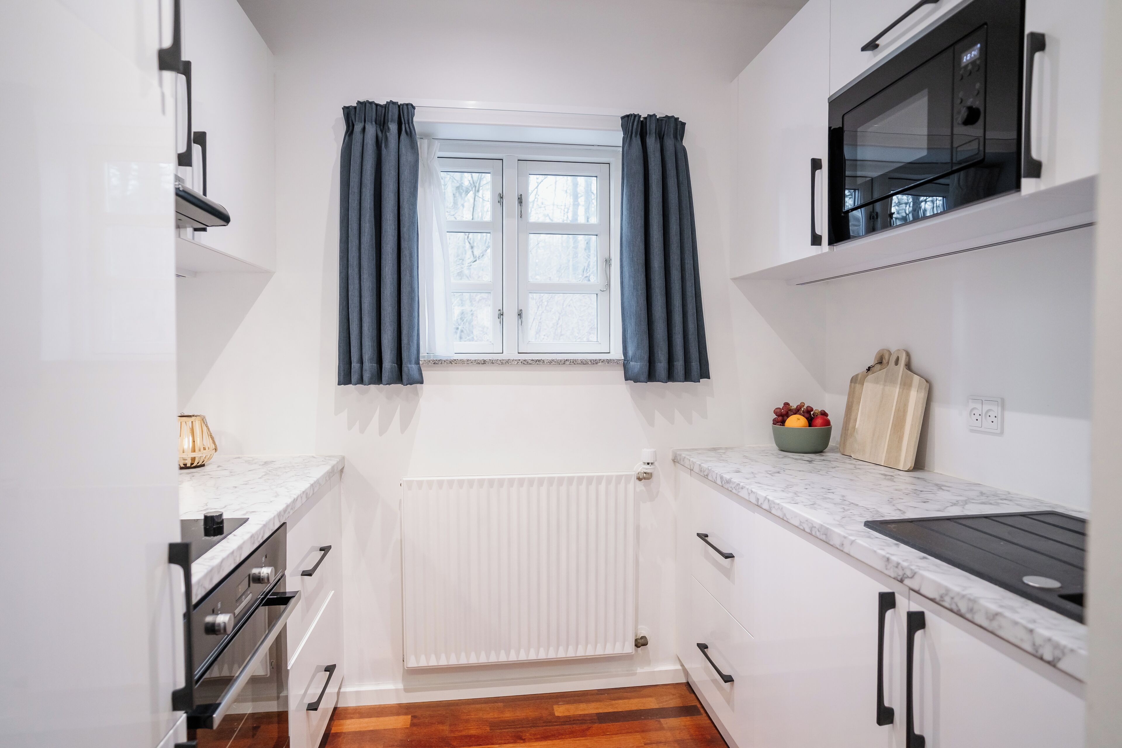 Uma cozinha compacta com armários brancos, forno embutido, micro-ondas e janela com cortinas.