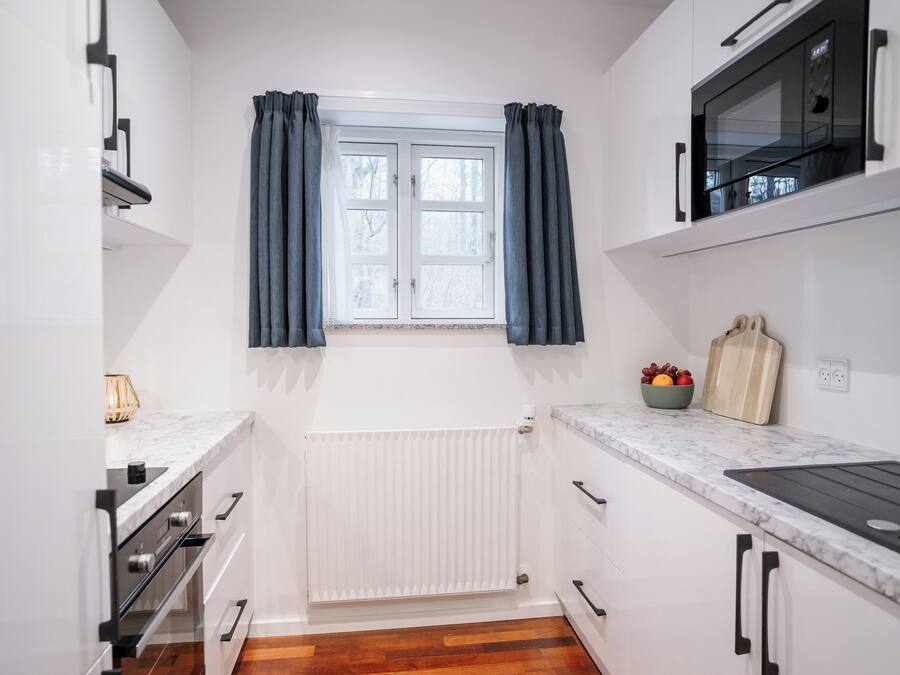 Una cocina compacta con armarios blancos, horno empotrado, microondas y ventana con cortinas.