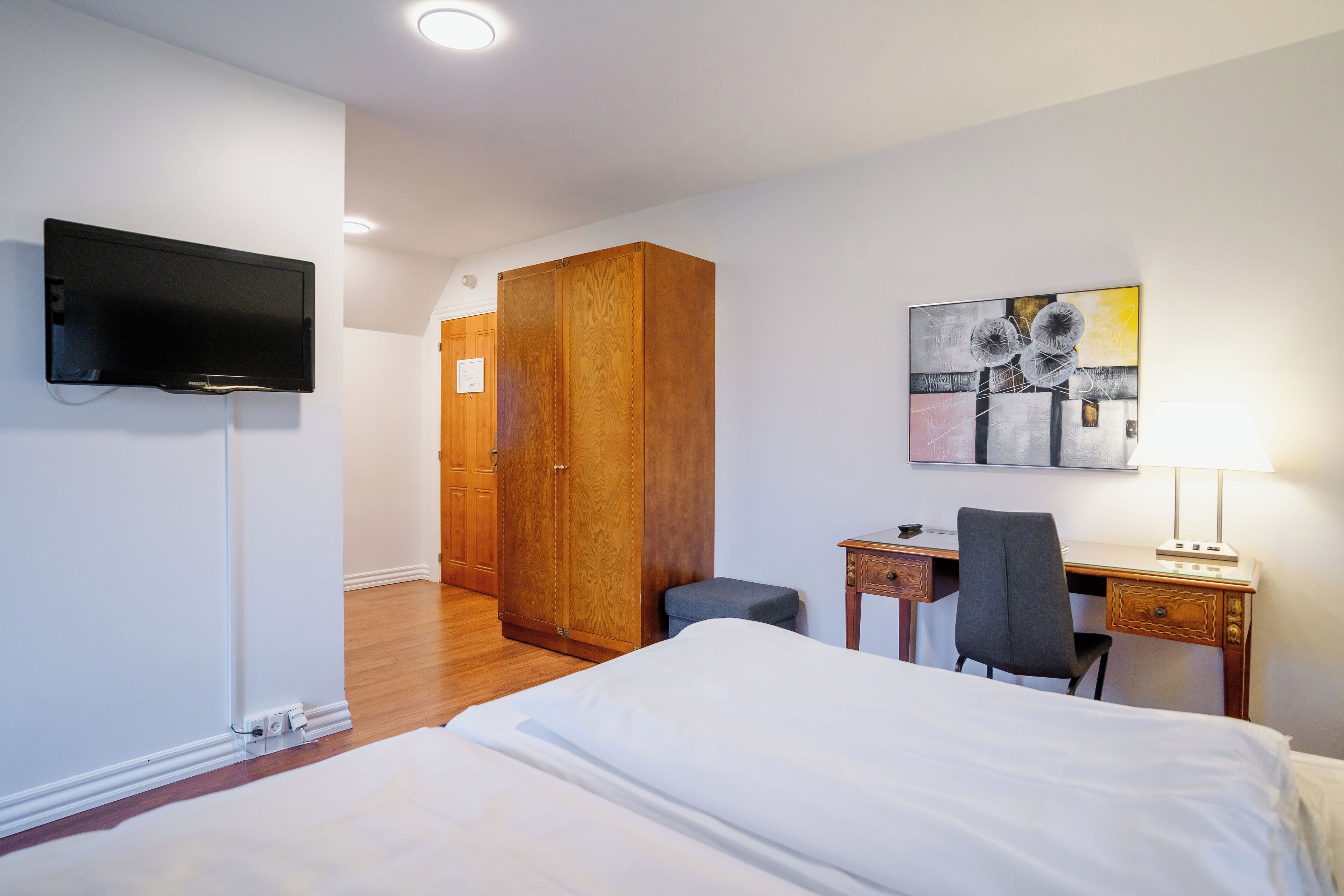 A hotel room with a bed, desk, chair, TV, wardrobe, and artwork on the wall.