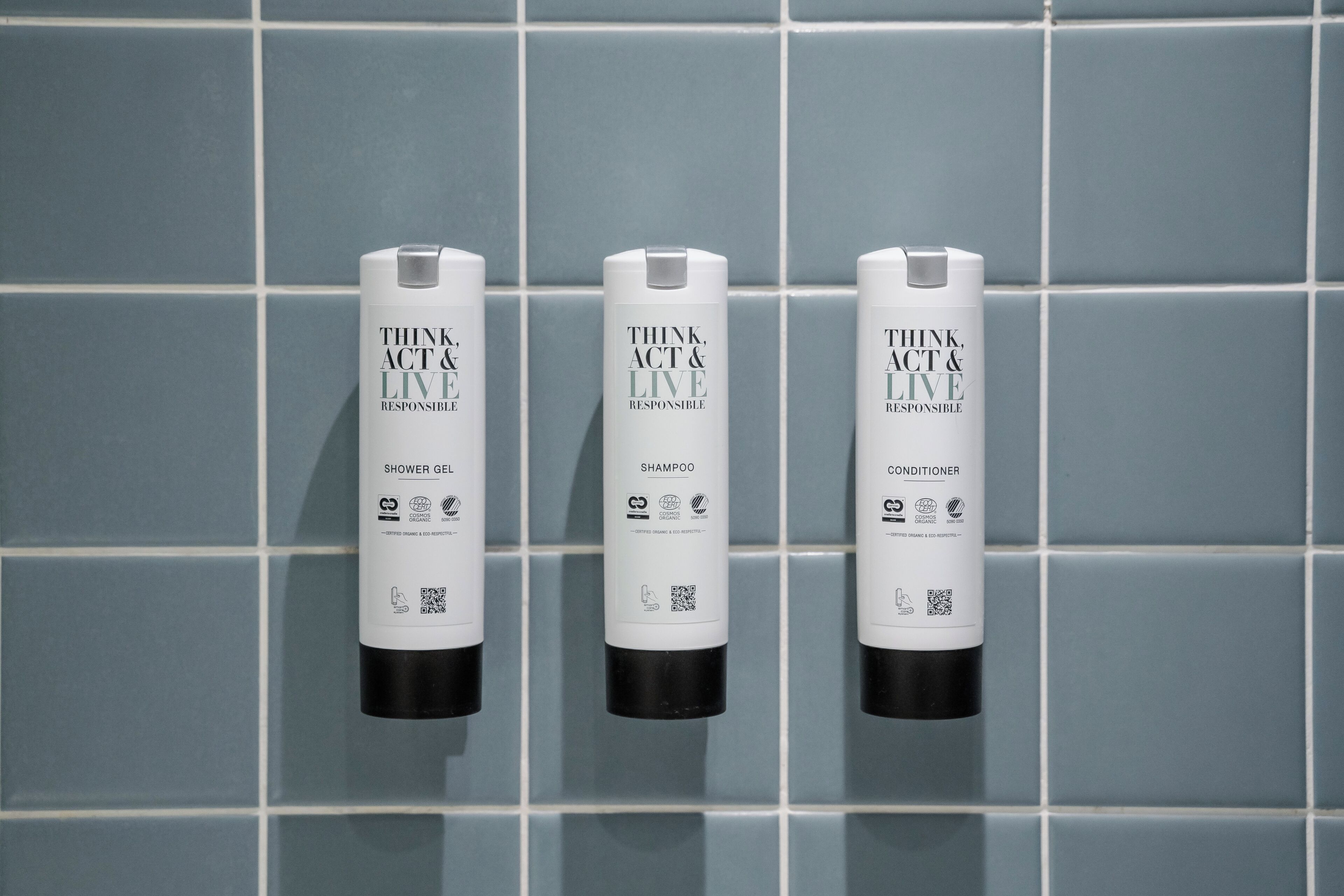 Three bottles of THINK ACT & LIVE products: SHOWER GEL, SHAMPOO, and CONDITIONER, arranged on a tiled wall.