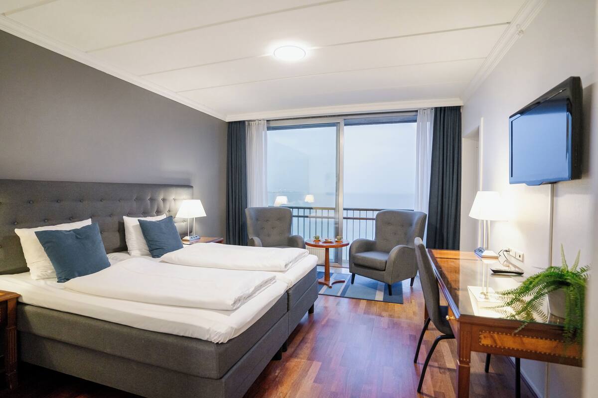 A hotel room with two beds, a desk with a chair, a TV, and a balcony with a view.