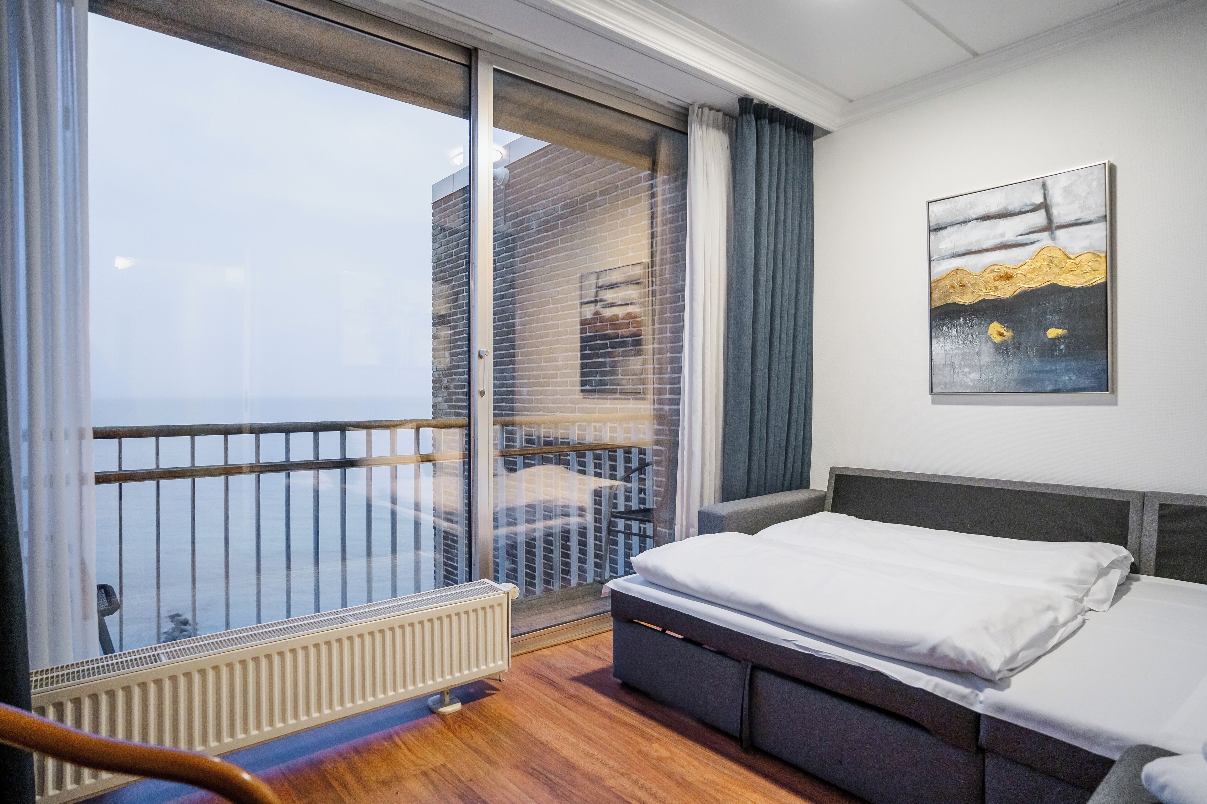 A hotel room with a large sliding glass door to a balcony, a bed with white bedding, a radiator, and a painting on the wall.
