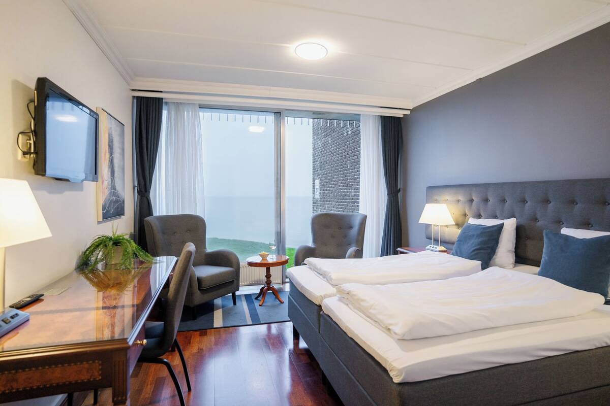 A hotel room with a large bed, two armchairs, a desk with a lamp, a TV, and a view of the outdoors.