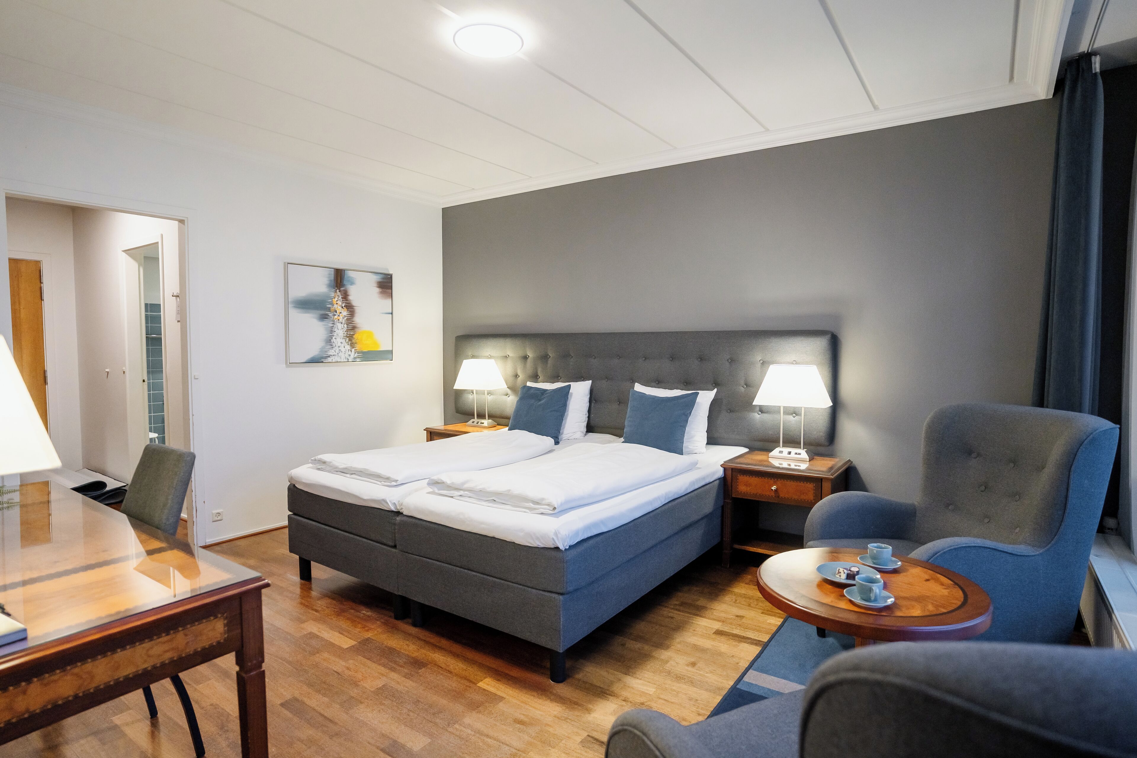 A hotel room with a double bed, two bedside tables with lamps, a small round table with a chair, and a desk with a chair.