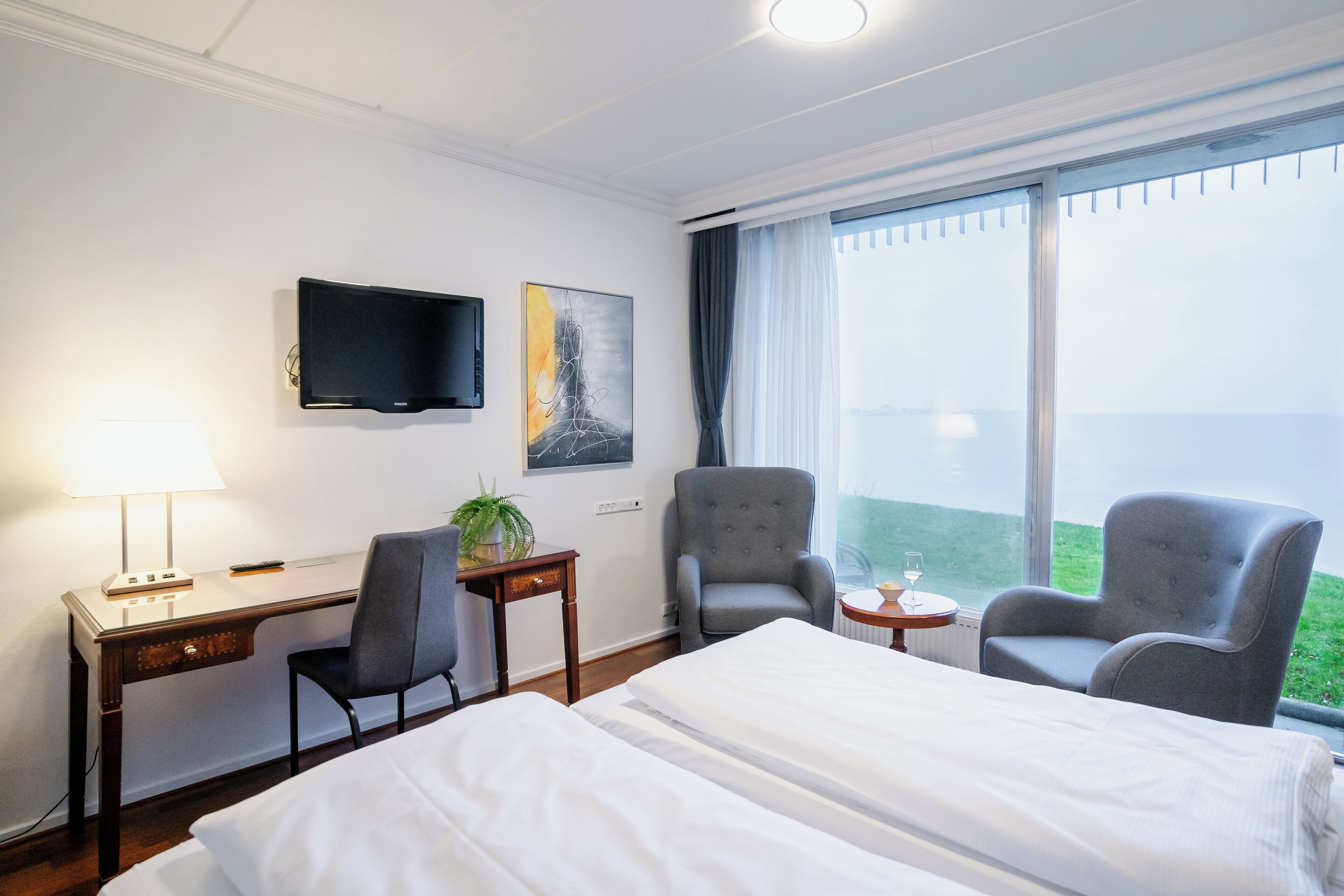 A hotel room with a bed, two armchairs, a desk with a lamp, a television, and a window with a view.