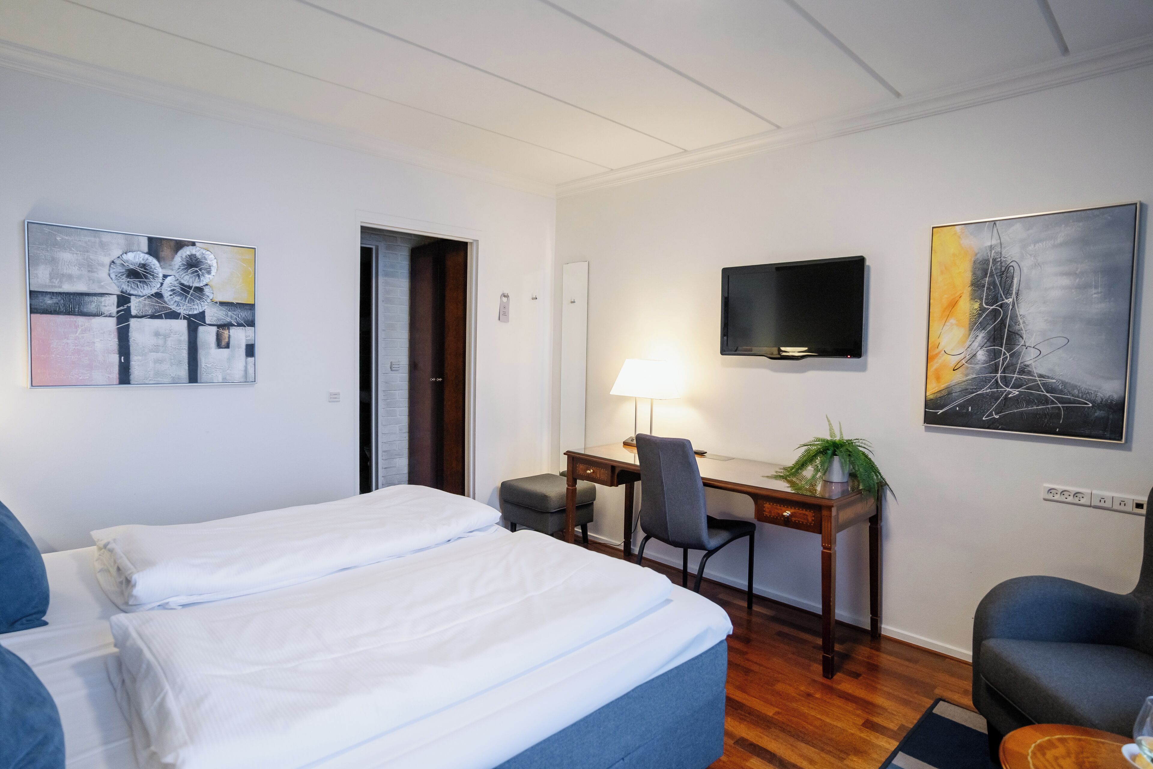 Habitación de hotel con dos camas, un escritorio, una silla, un televisor y obras de arte en las paredes.