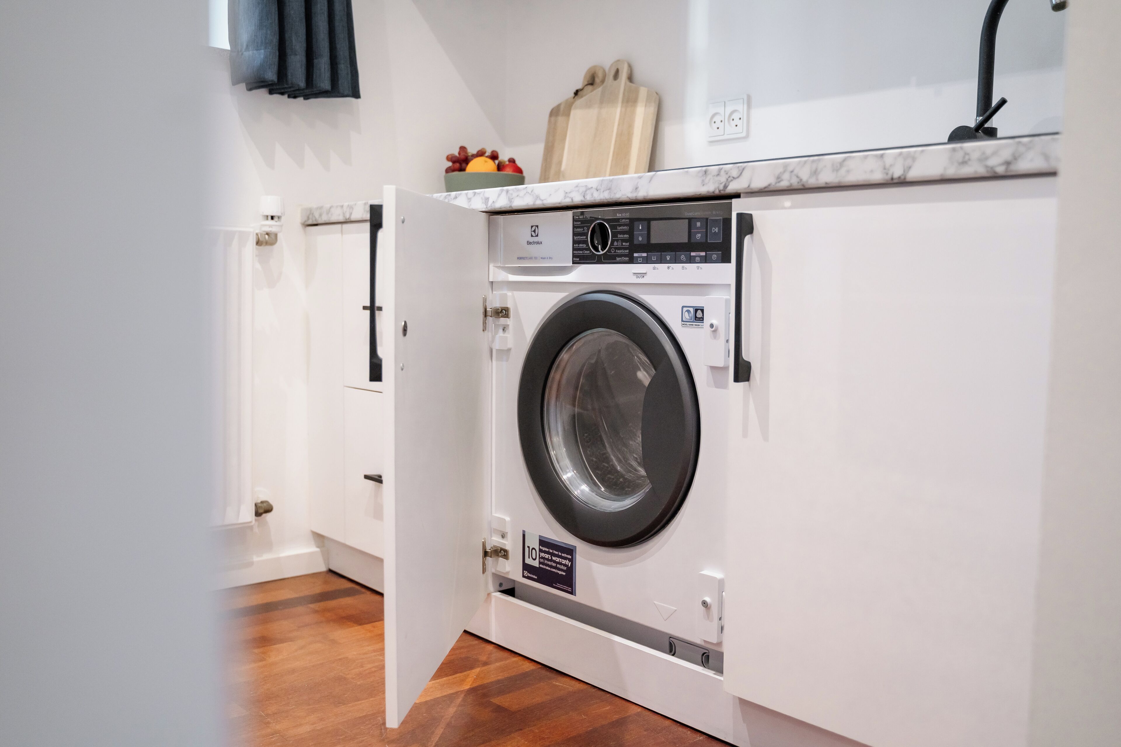 A compact, modern laundry room with a built-in washer and dryer, a wooden cutting board, and a fruit bowl on the countertop.