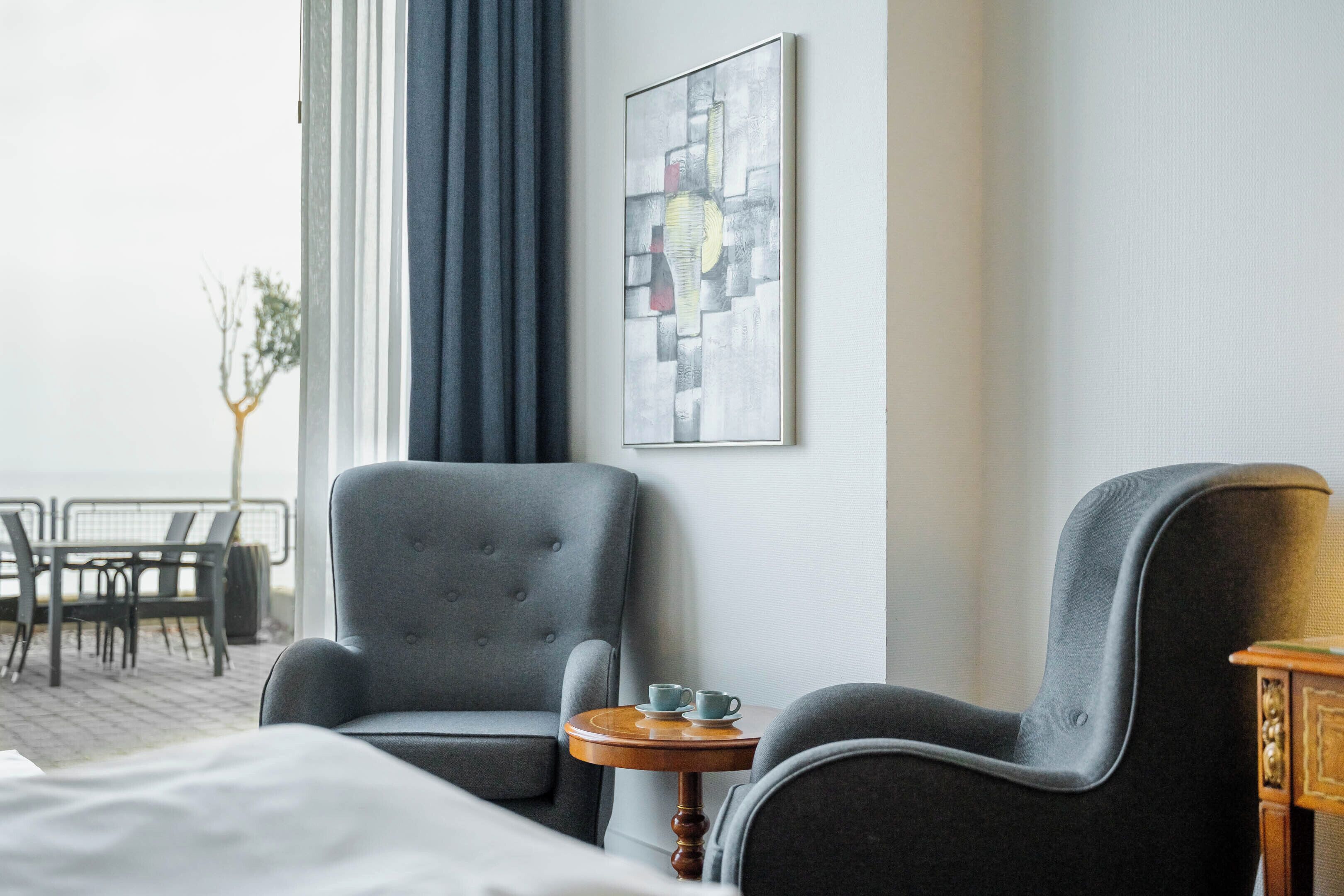 A hotel room with a bed, two grey armchairs, a small wooden side table with two cups, a framed abstract painting on the wall, and a view of the outdoors through a window.