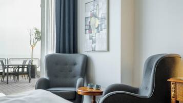 Habitación de hotel con una cama, dos sillones grises, una mesita de madera con dos tazas, un cuadro abstracto enmarcado en la pared y vistas al exterior a través de una ventana.