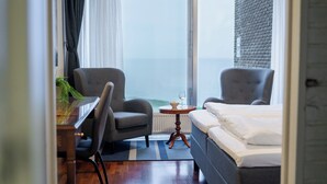 Un cuarto de hotel con cama, dos sillones, una mesita con una copa de vino y vista al mar a través de un ventanal grande.