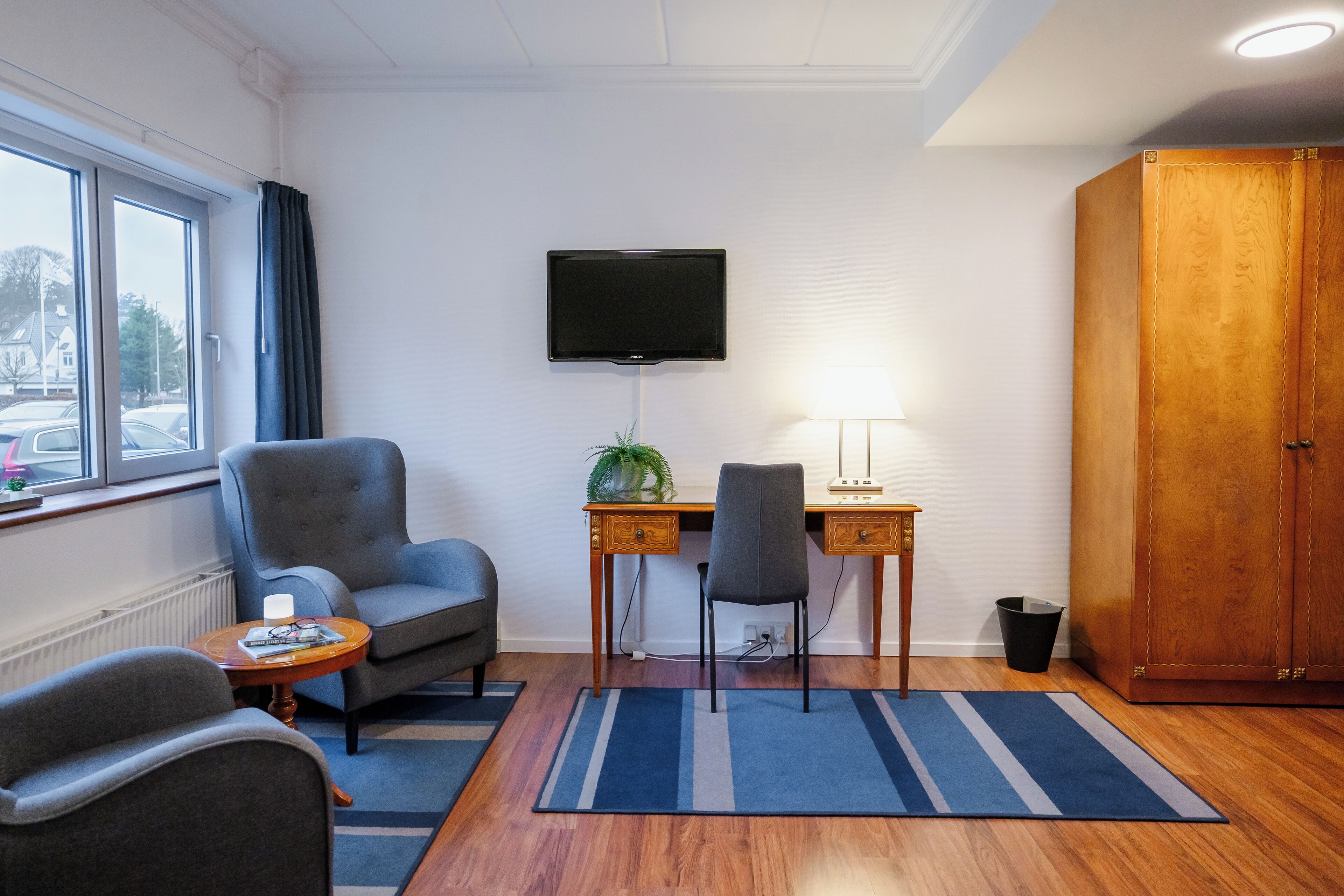 A room with a desk, two chairs, a TV mounted on the wall, a wooden wardrobe, and a window with curtains.