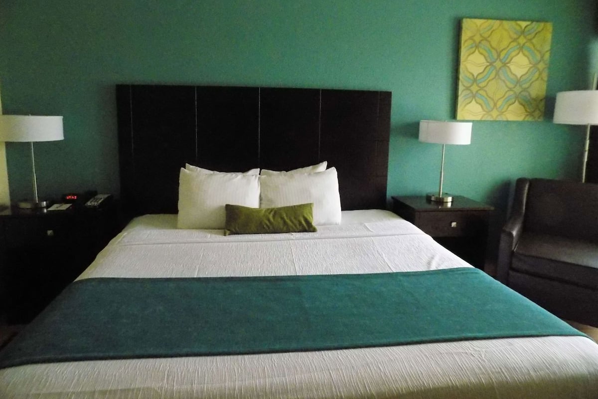 Una habitación de hotel con una cama, dos mesitas de noche, una silla y una pared con un cuadro de motivos geométricos.