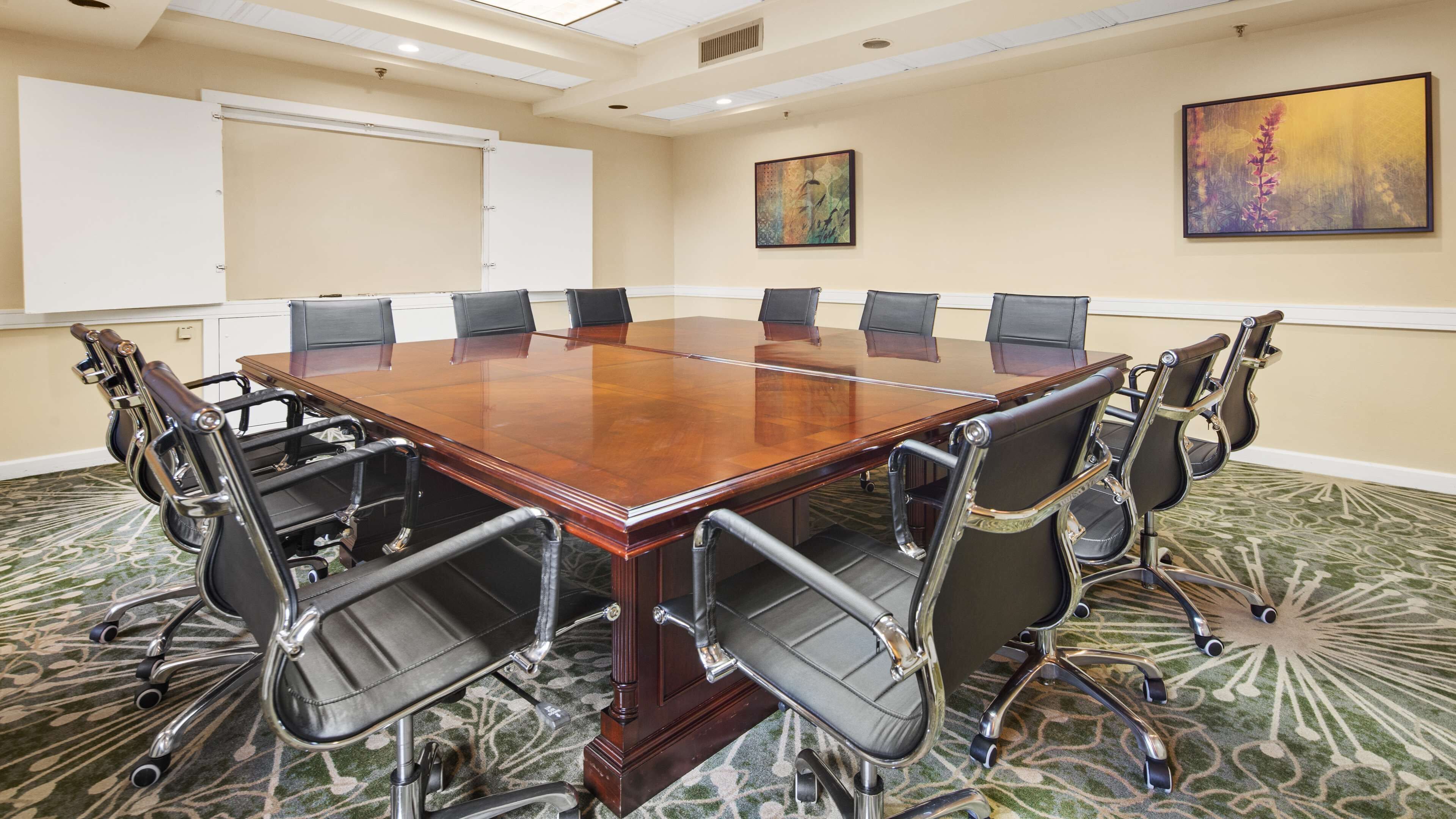 A conference room with a large wooden table, eight black chairs with metal frames, a whiteboard, and two paintings on the walls.