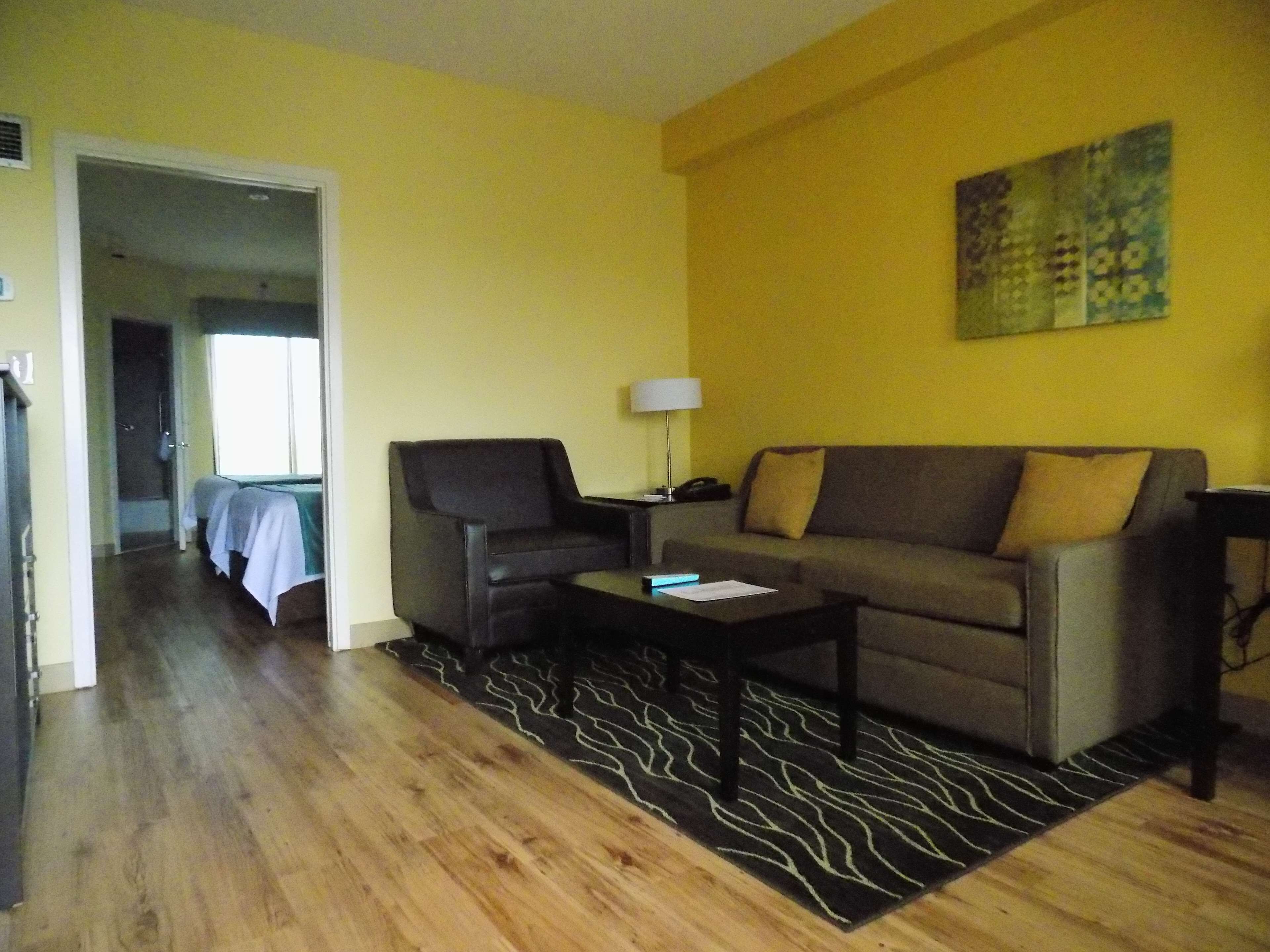 A living room with a sofa, armchair, and coffee table. There is a painting on the wall and a door leading to another room.