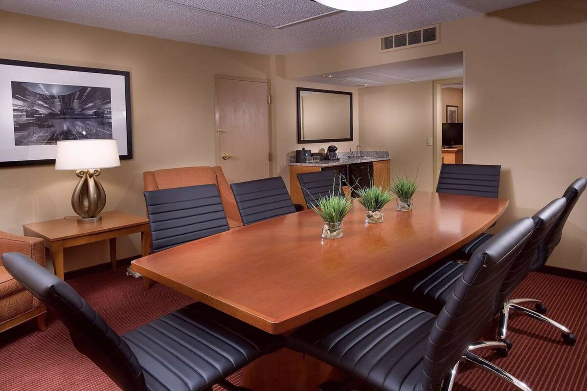 Une salle de conférence avec une table en bois, des fauteuils en cuir noir, une petite table d’appoint avec une lampe et des plantes en pot.
