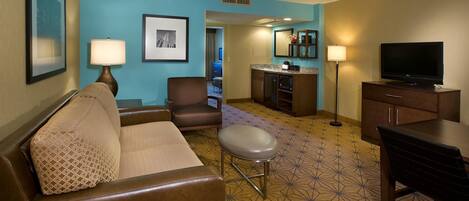 Habitación de hotel con sofá, sillón, reposapiés, televisor de pantalla plana, escritorio, lámpara, microondas y una pequeña kitchenette.