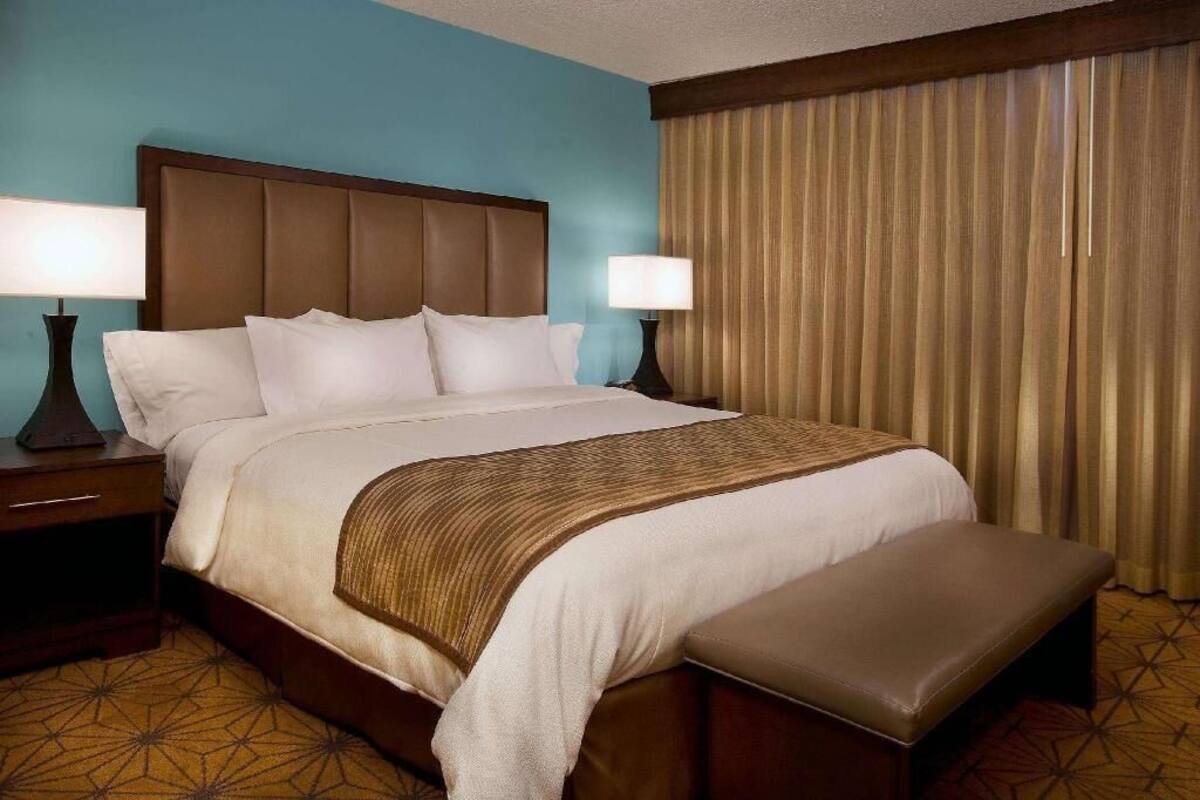 Une chambre d’hôtel avec un grand lit, deux tables de chevet avec des lampes, un banc au pied du lit et une fenêtre avec des rideaux.