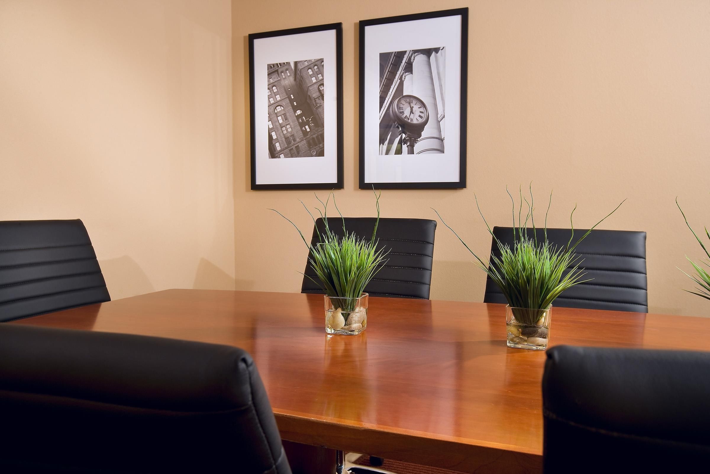 A conference room with a wooden table, four black chairs, two framed black and white photographs on the wall, and two small potted plants with green foliage.