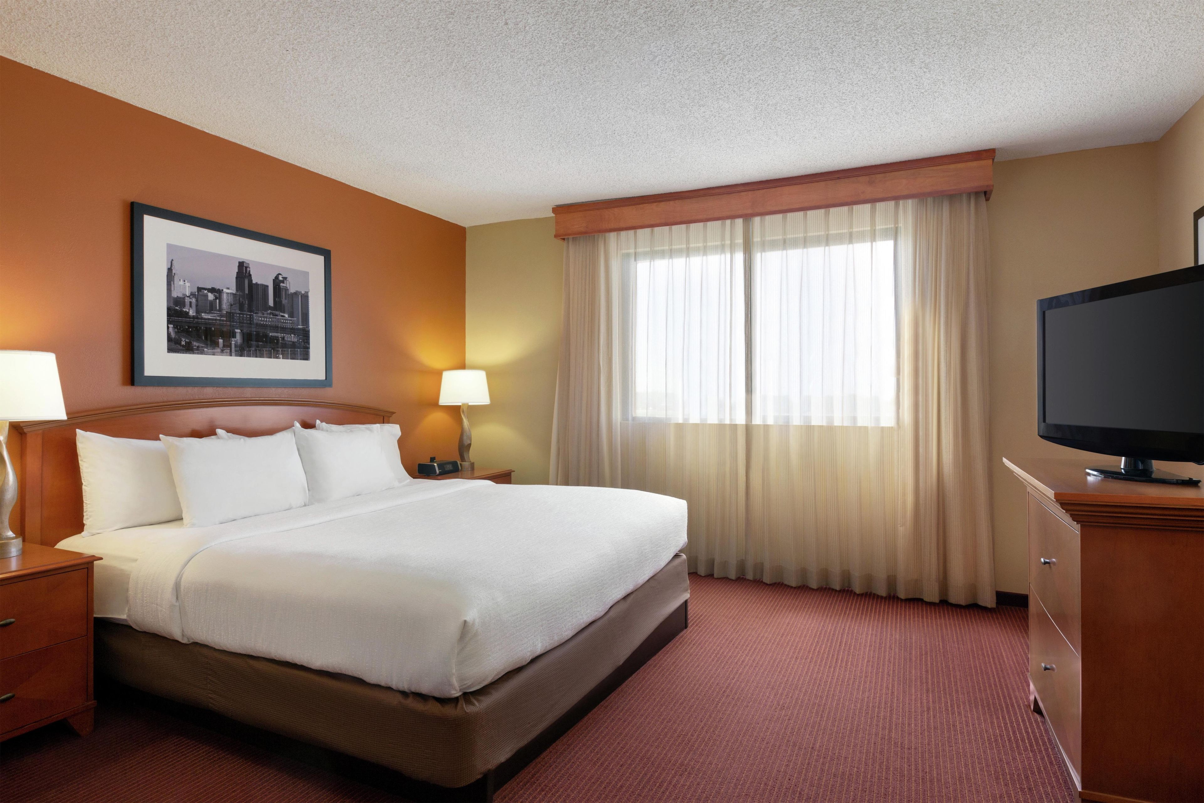 A hotel room with a large bed, a television, a nightstand, a lamp, a framed cityscape picture, and a window with curtains.