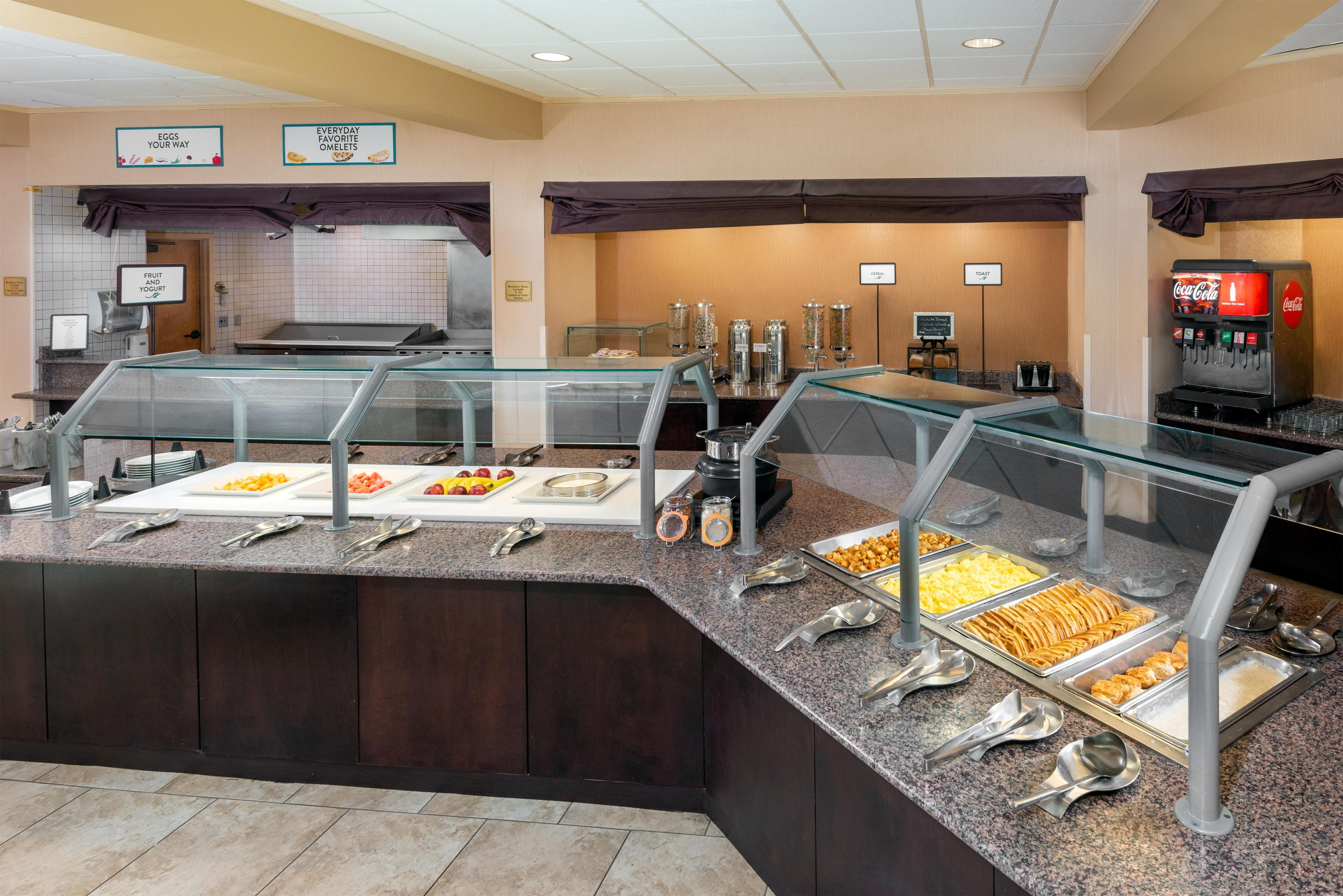 Zona de buffet con diversas opciones de comida, incluyendo frutas, cereales y un dispensador de Coca-Cola.