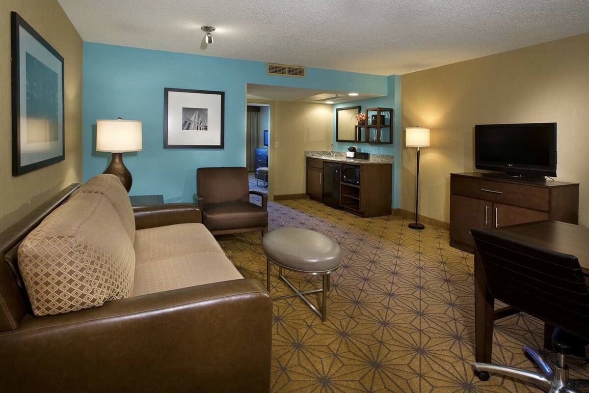Une chambre d’hôtel avec un canapé en cuir marron, un petit tabouret, un bureau avec une chaise, une télévision, un coin cuisine et une salle de bain visible à travers une porte ouverte.