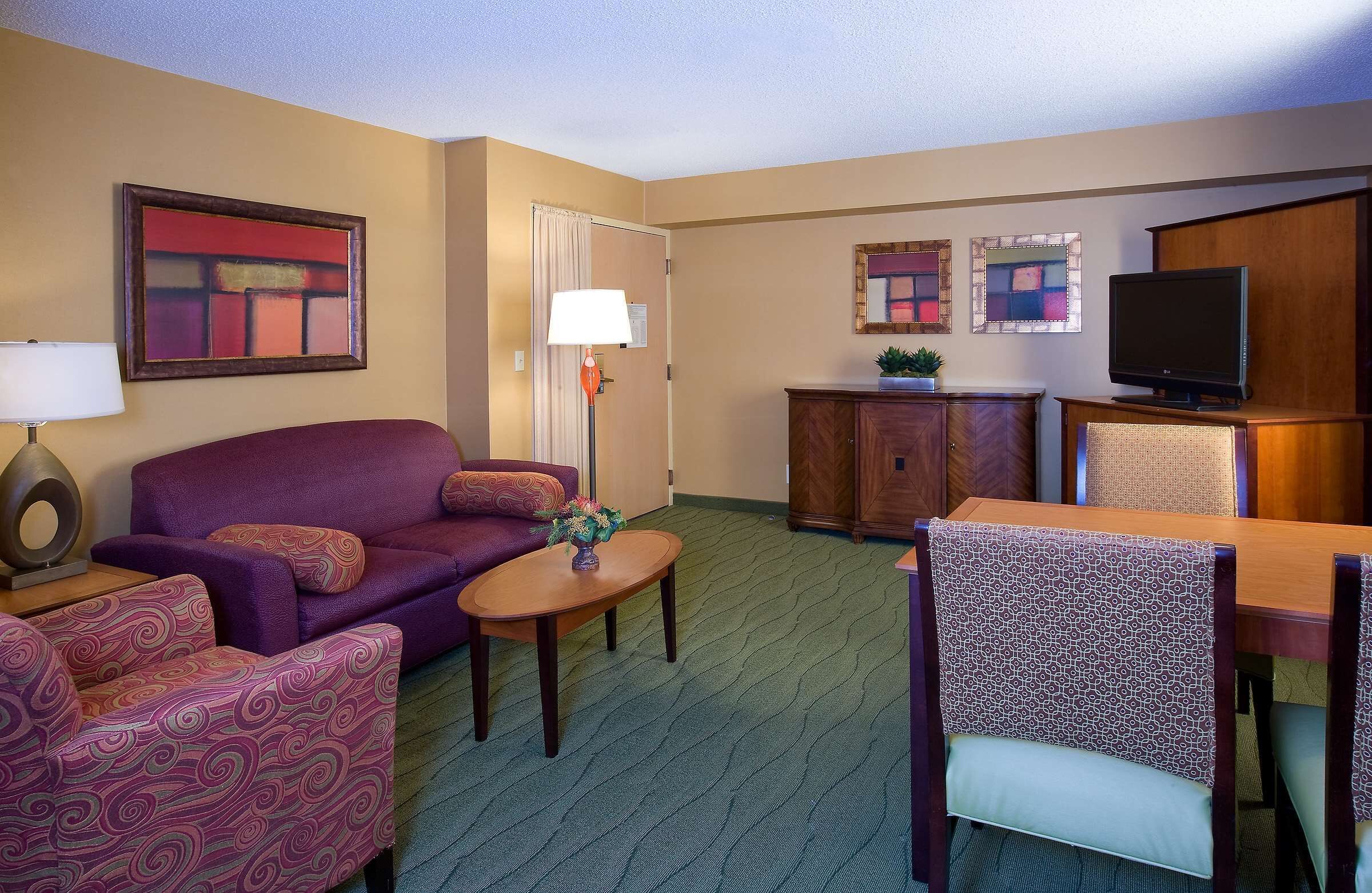 A hotel room with a purple sofa, a small table with a vase, a wooden cabinet, a television, and a dining area with a patterned chair.