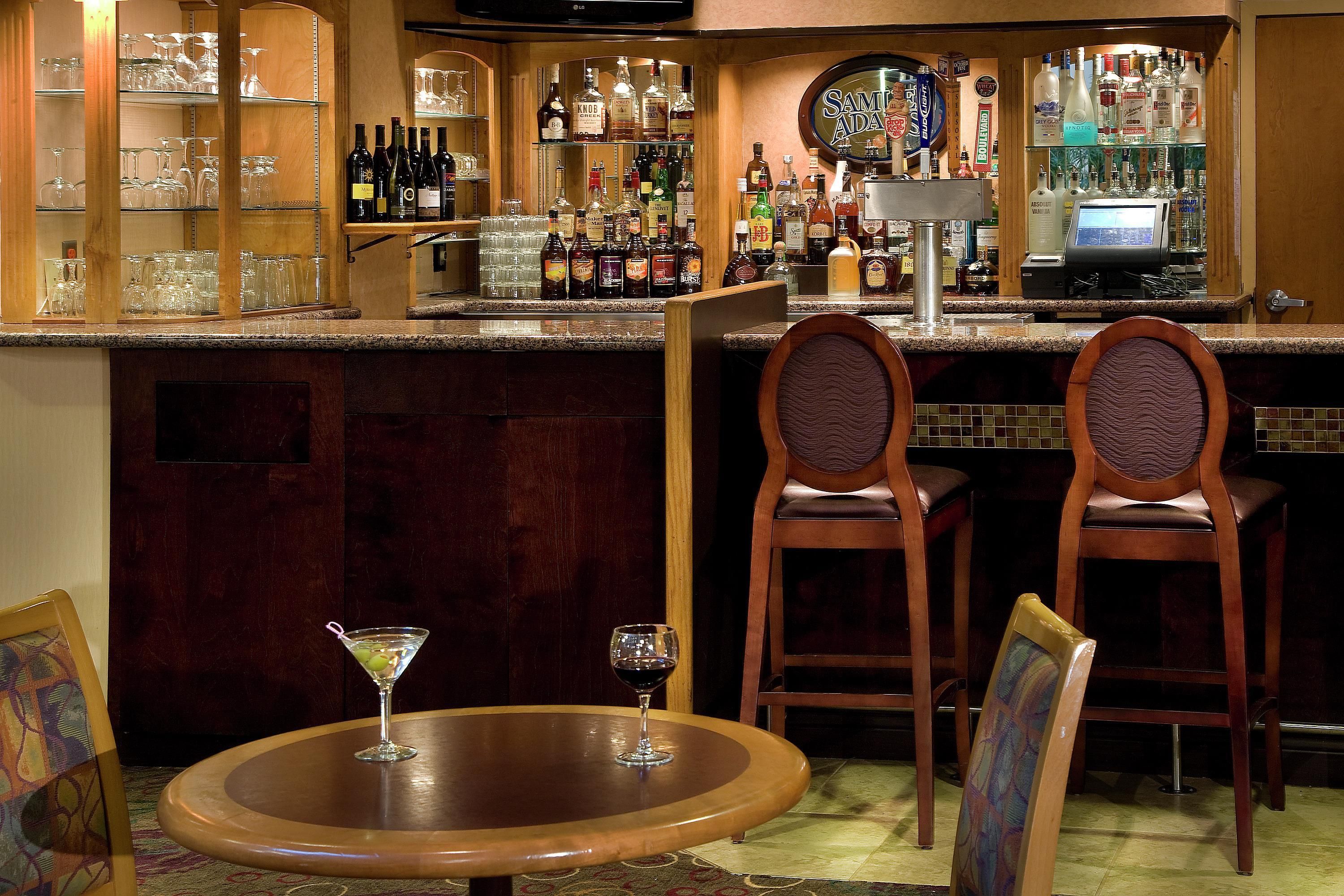 Un coin bar avec deux chaises en bois, une table ronde sur laquelle se trouve un verre à martini, et diverses bouteilles d’alcool disposées sur des étagères.