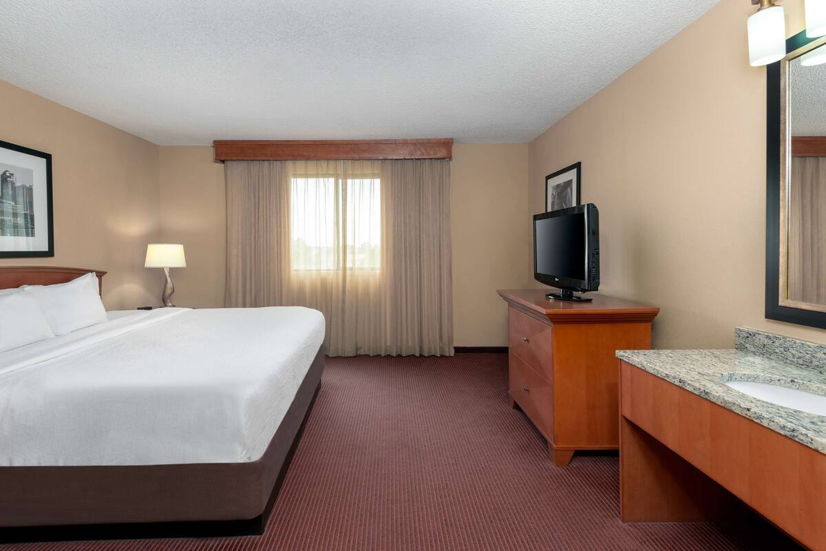 Une chambre d’hôtel avec un grand lit, un téléviseur posé sur une commode en bois, une salle de bain avec un plan de toilette en granit et une fenêtre avec des rideaux.