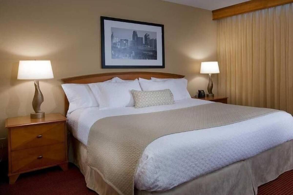 Une chambre d’hôtel avec un grand lit, deux tables de chevet, un tableau encadré représentant une vue sur la ville accroché au mur, et une fenêtre avec des rideaux.