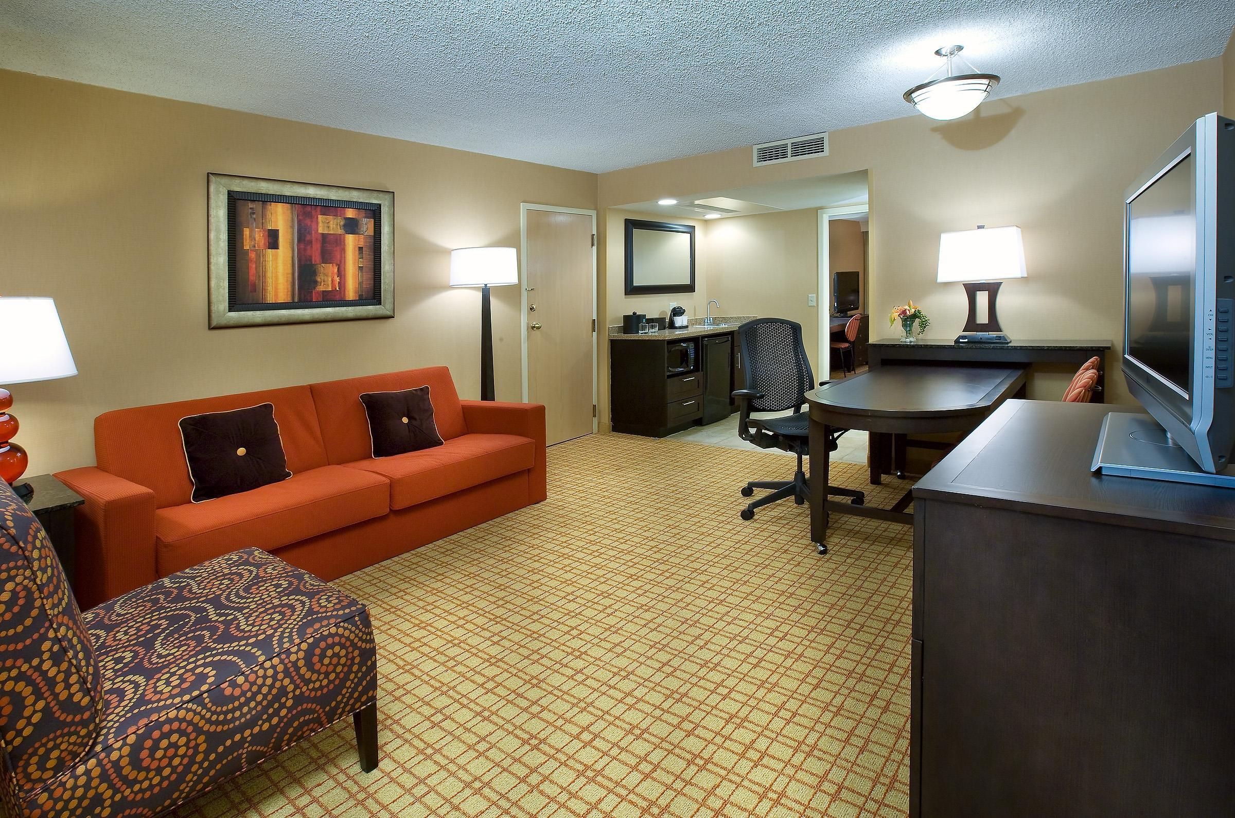 A hotel room with a sofa, armchair, desk, television, and a painting on the wall.