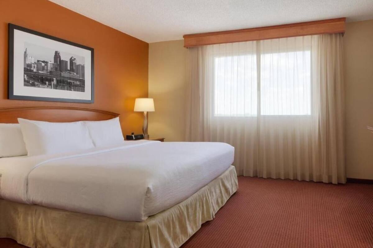 Une chambre d’hôtel avec un grand lit, une tête de lit, une table de chevet, une lampe, un tableau encadré au mur et une fenêtre avec des rideaux.