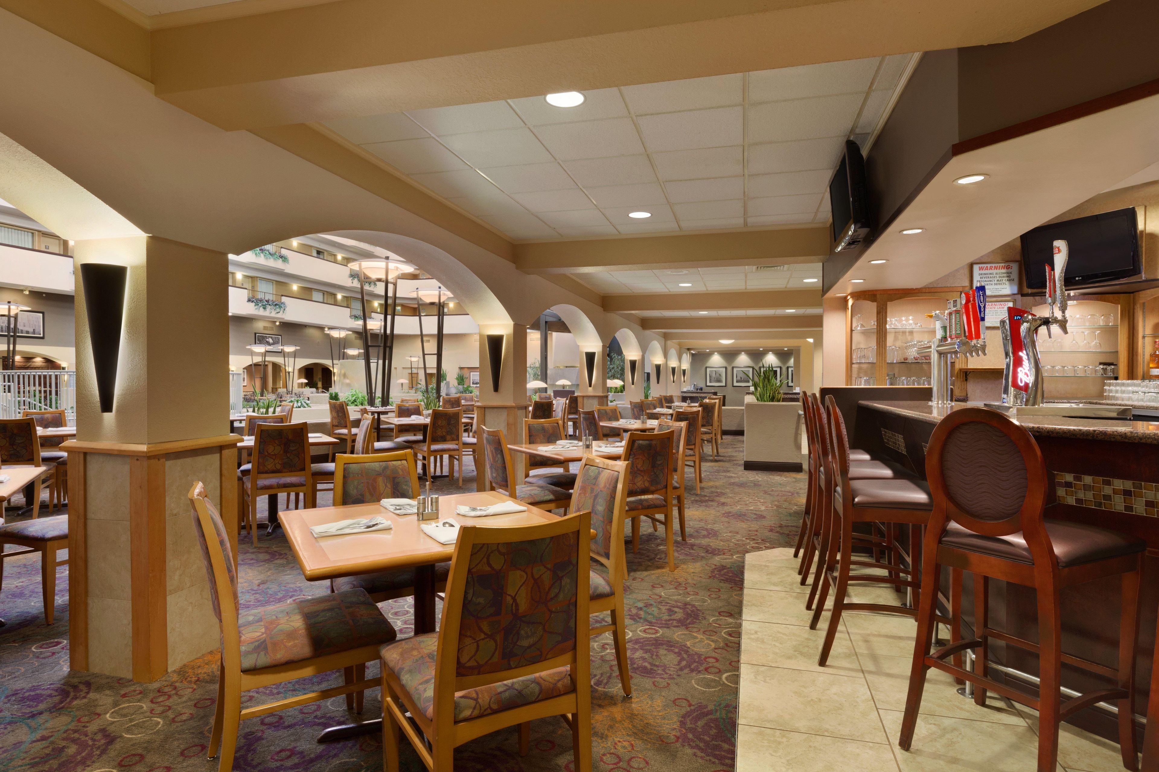 A hotel dining area with multiple tables and chairs, a bar area with a television, and a view of the hotel atrium.