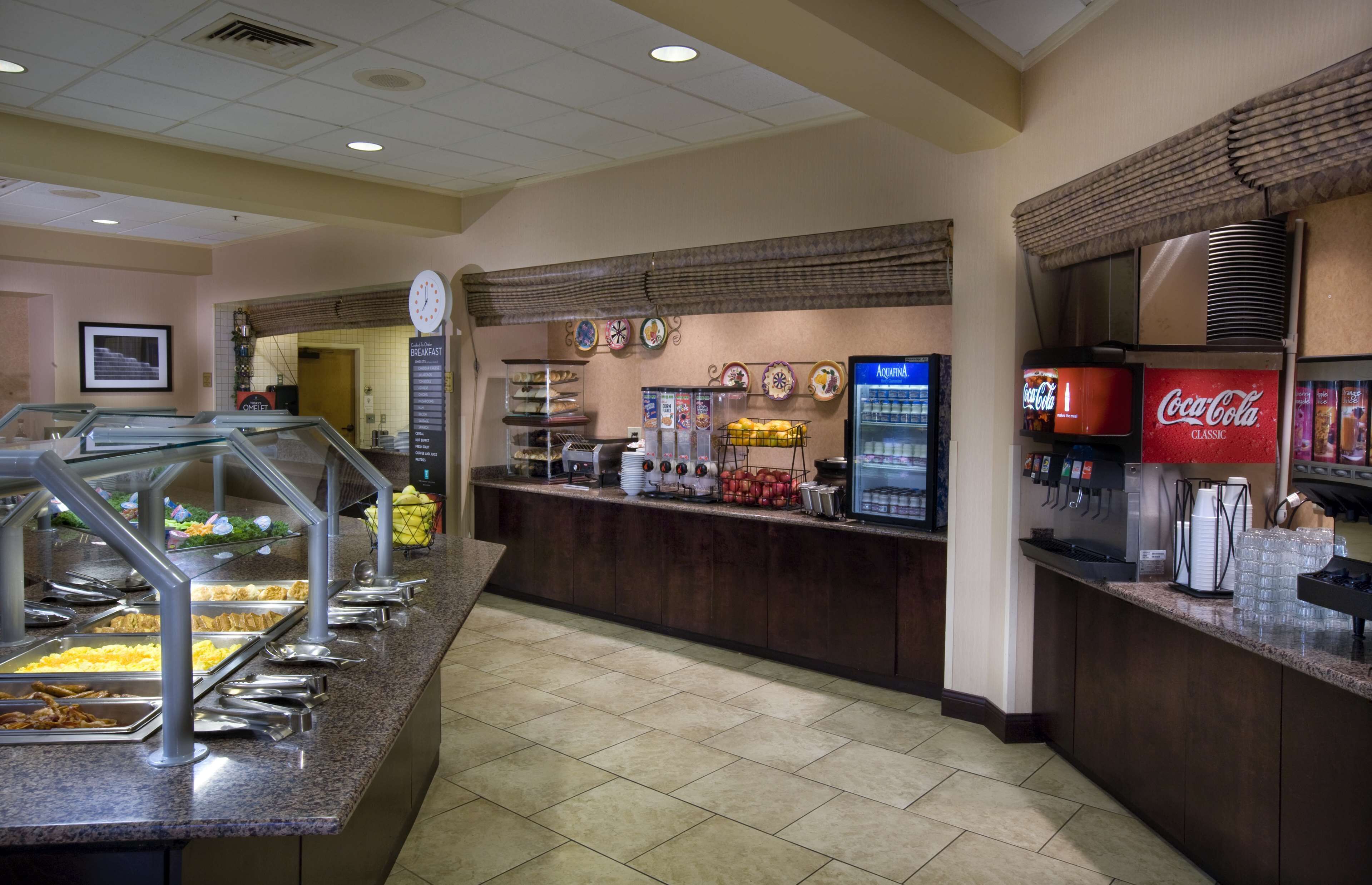 Uma área de buffet com diversas estações de comida, um refrigerador Coca-Cola e um relógio na parede.