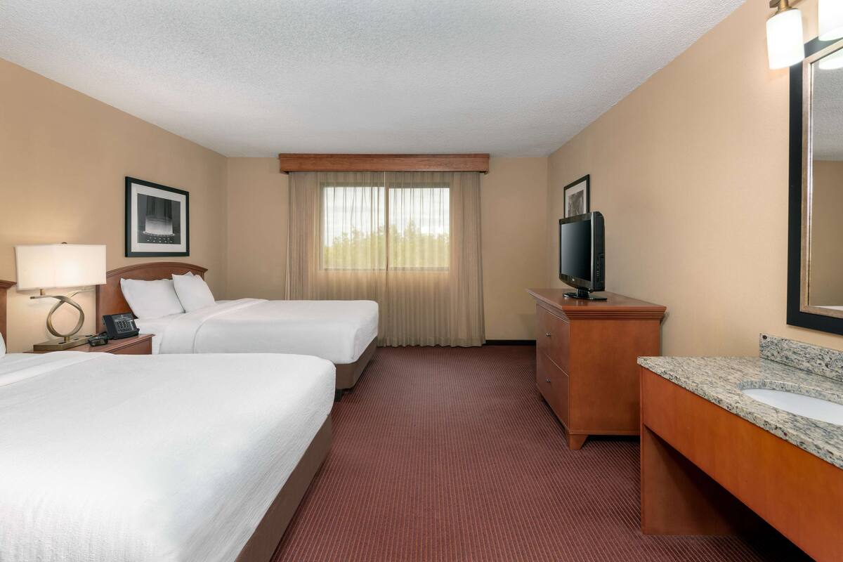 Une chambre d’hôtel avec deux lits, une commode en bois, une télévision et une grande fenêtre avec des rideaux.
