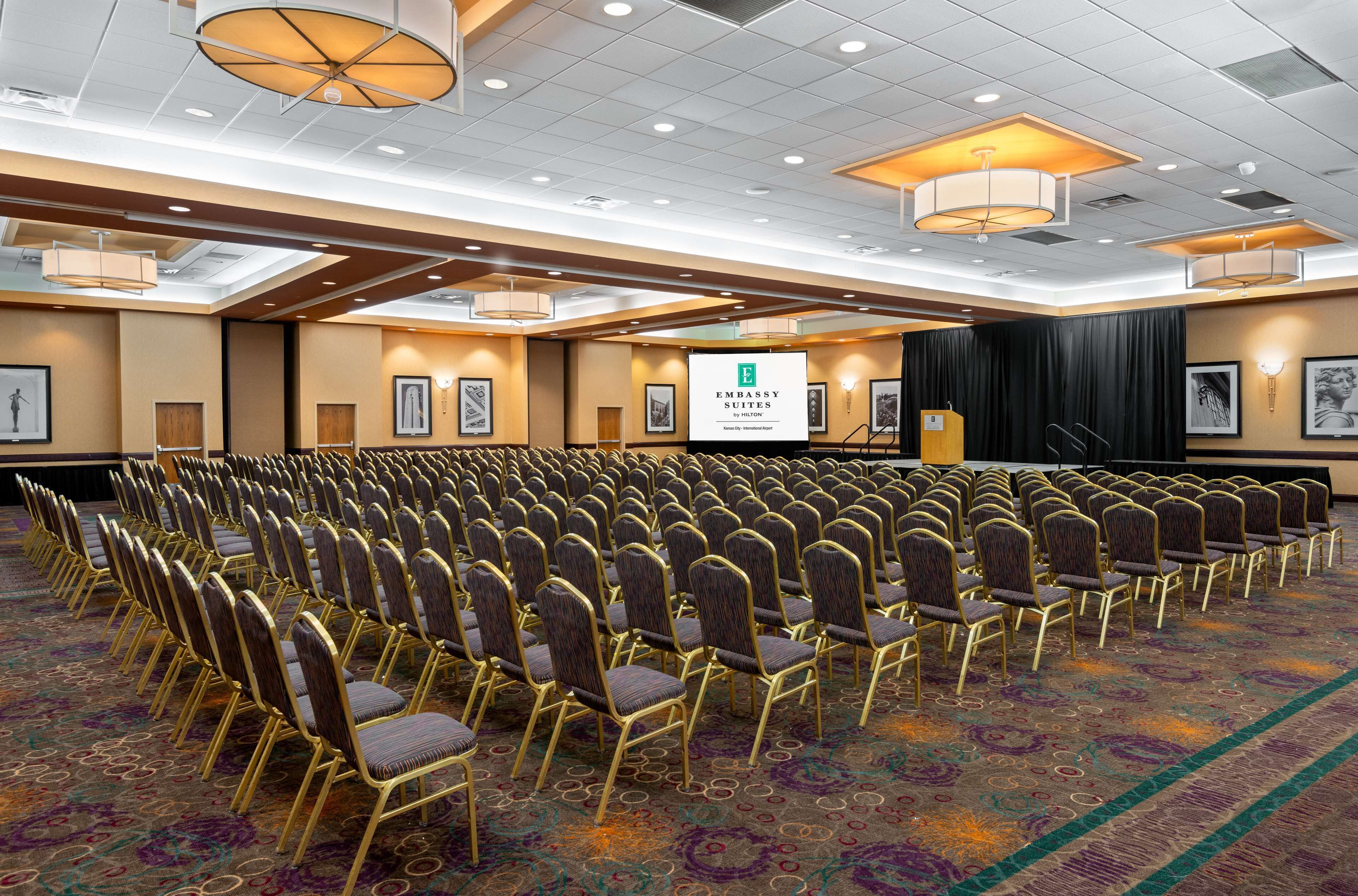 Een congreszaal met rijen stoelen, een spreekgestalte en een banner met de tekst 'Embassy Suites by Hilton'.