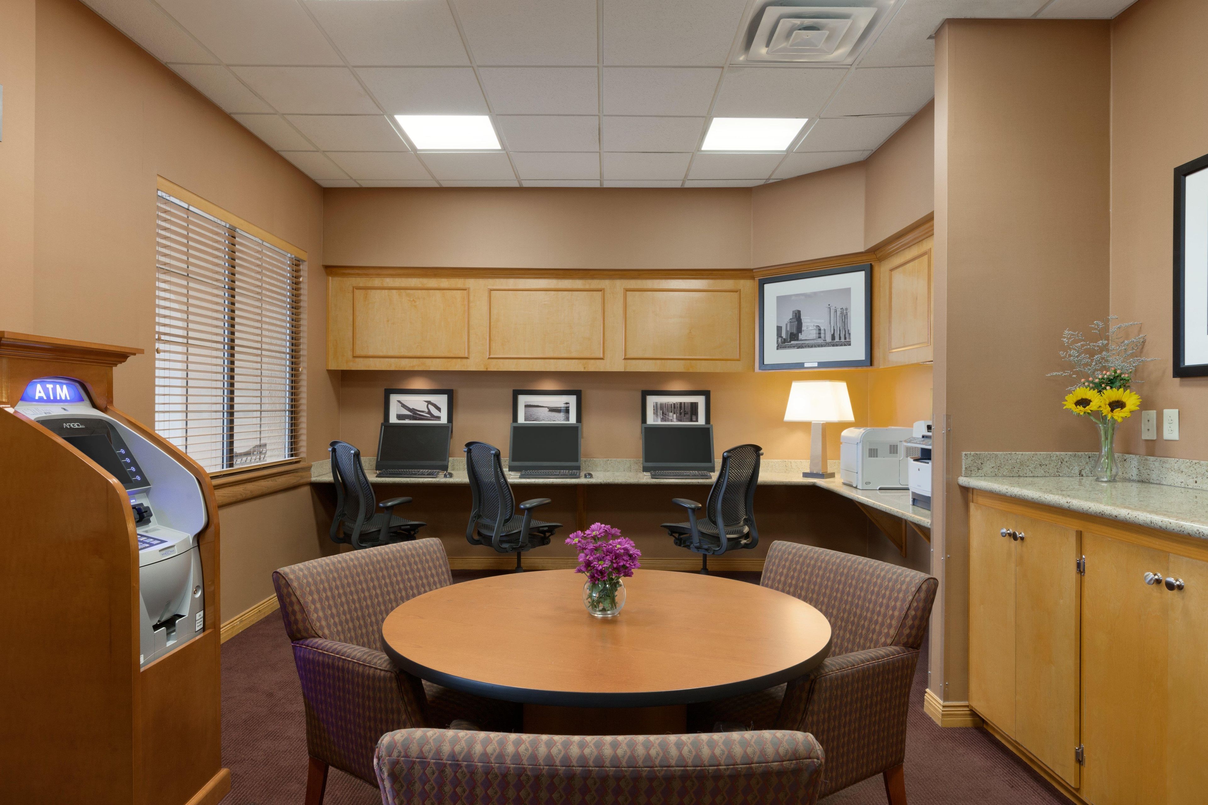 A conference room with a round table, four chairs, a desk with a computer, an ATM, and framed pictures on the wall.