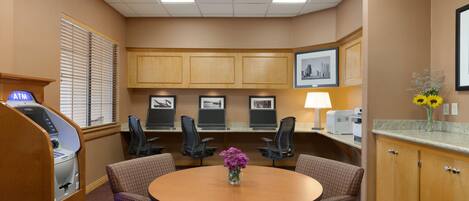 A conference room with a round table, four chairs, a desk with a computer, an ATM, and framed pictures on the wall.