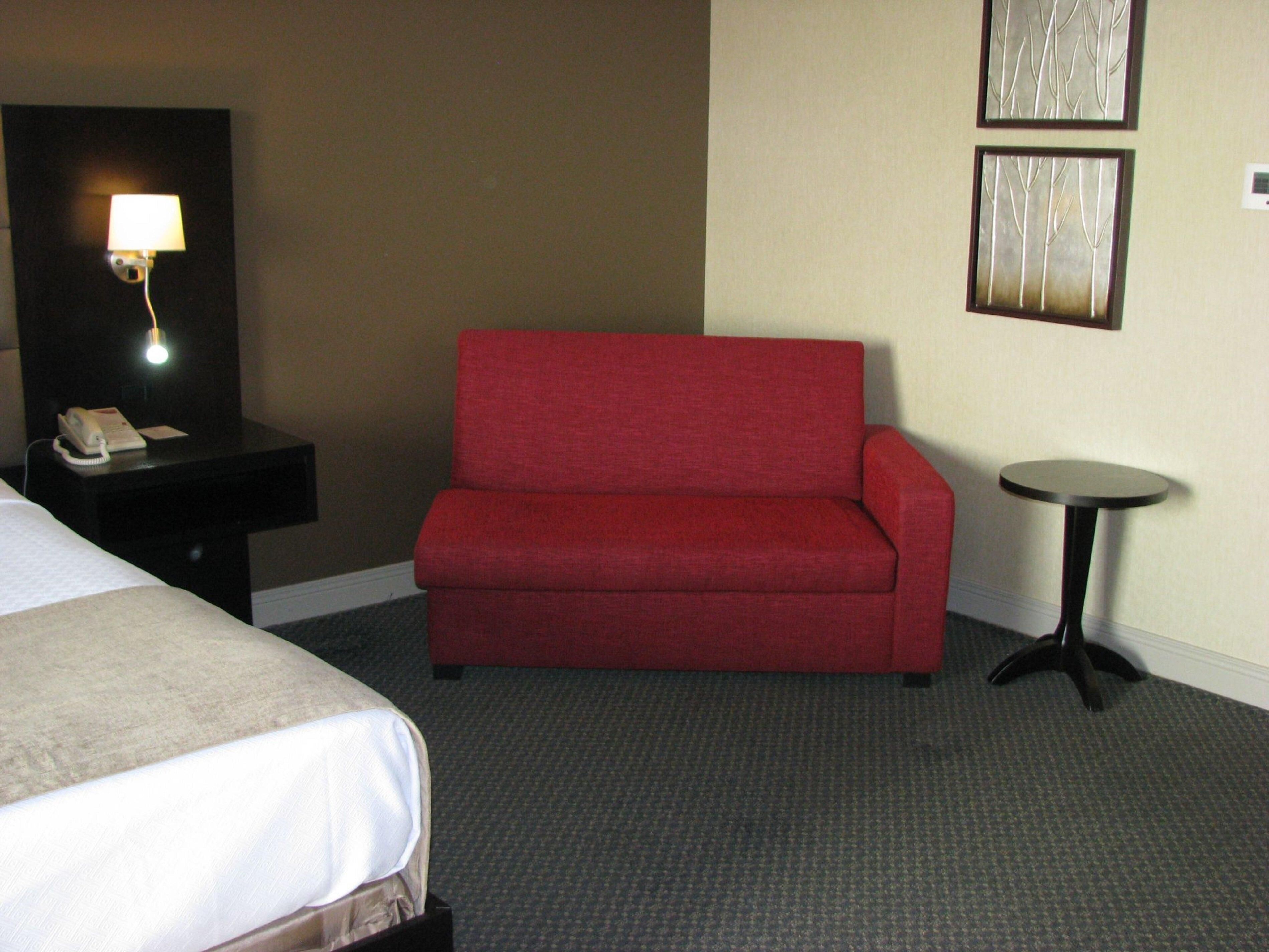 A hotel room with a red sofa, a small round table, a bedside table with a lamp, and a bed with a white comforter.