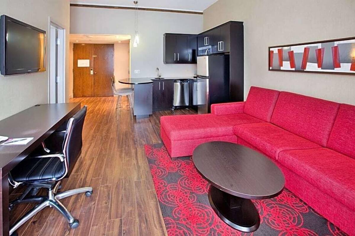 A modern hotel room with a red sofa, a dining table with chairs, a kitchen area, and a TV mounted on the wall.