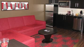 Un espacio habitable compacto con un sofá rojo, una mesita redonda pequeña, una zona de cocina con refrigerador y microondas, y una alfombra estampada.