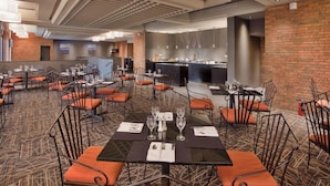 A dining area with tables set for four, each with a black tablecloth, white napkins, and silverware. Orange cushioned chairs surround the tables. A brick wall and a modern kitchen area are visible in the background. - Wyndham Gatineau-Ottawa & Conference Centre (Gatineau)