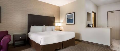 A hotel room with a large bed, a nightstand, a lamp, a chair, and a framed picture on the wall.