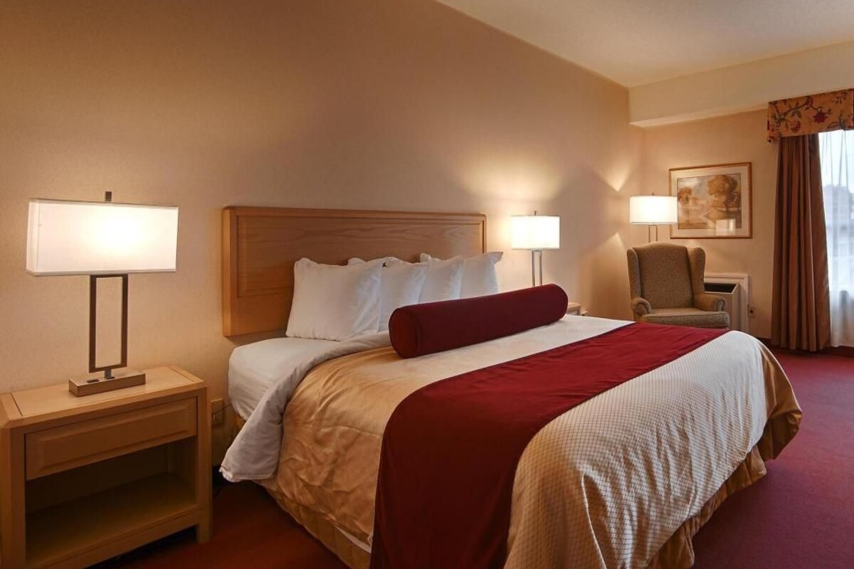 Habitación de hotel con cama, dos lámparas de noche, mesita de noche, una silla y un cuadro en la pared.