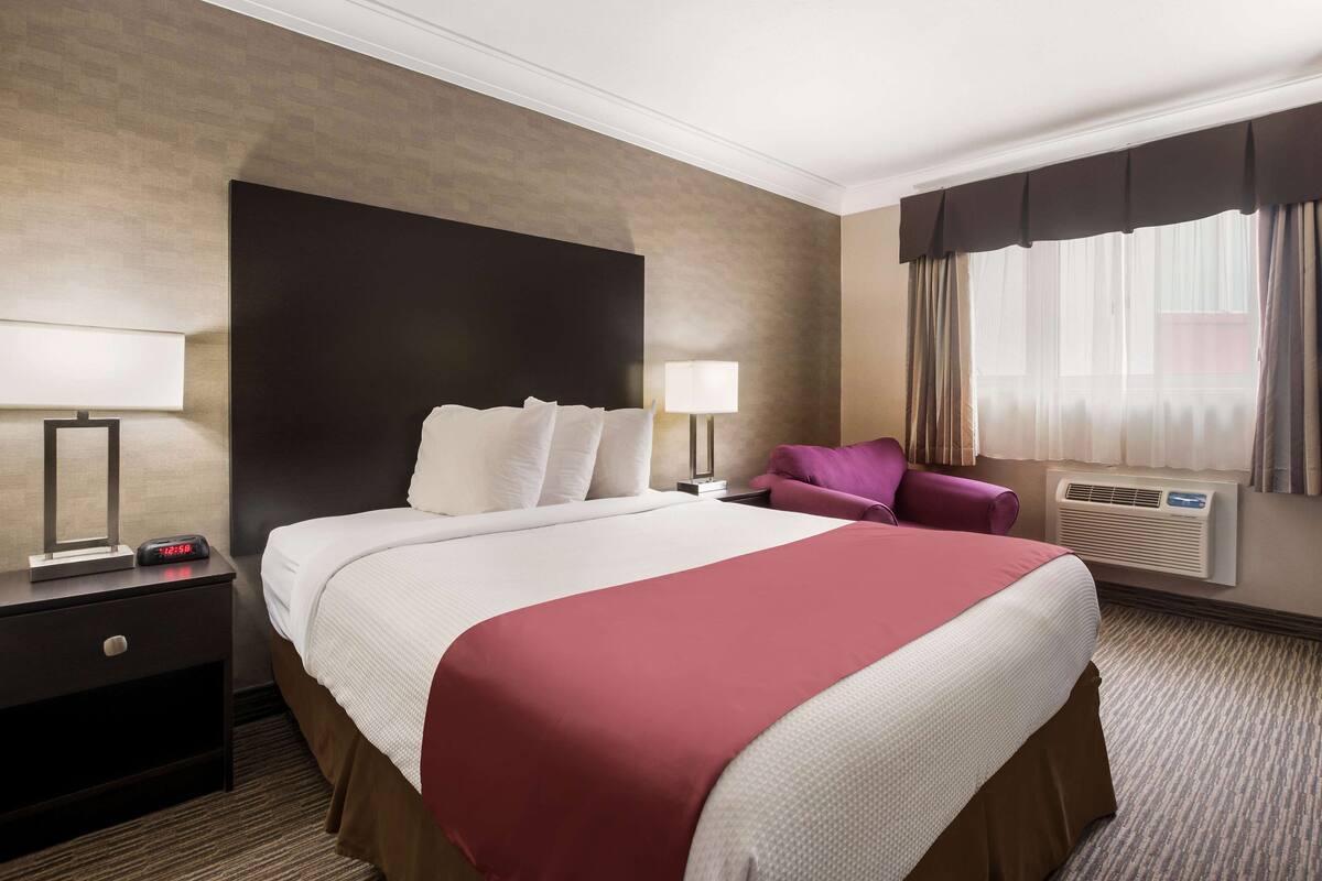 Habitación de hotel con una cama grande, una mesita de noche con lámpara, una silla y un ventanal con cortinas.
