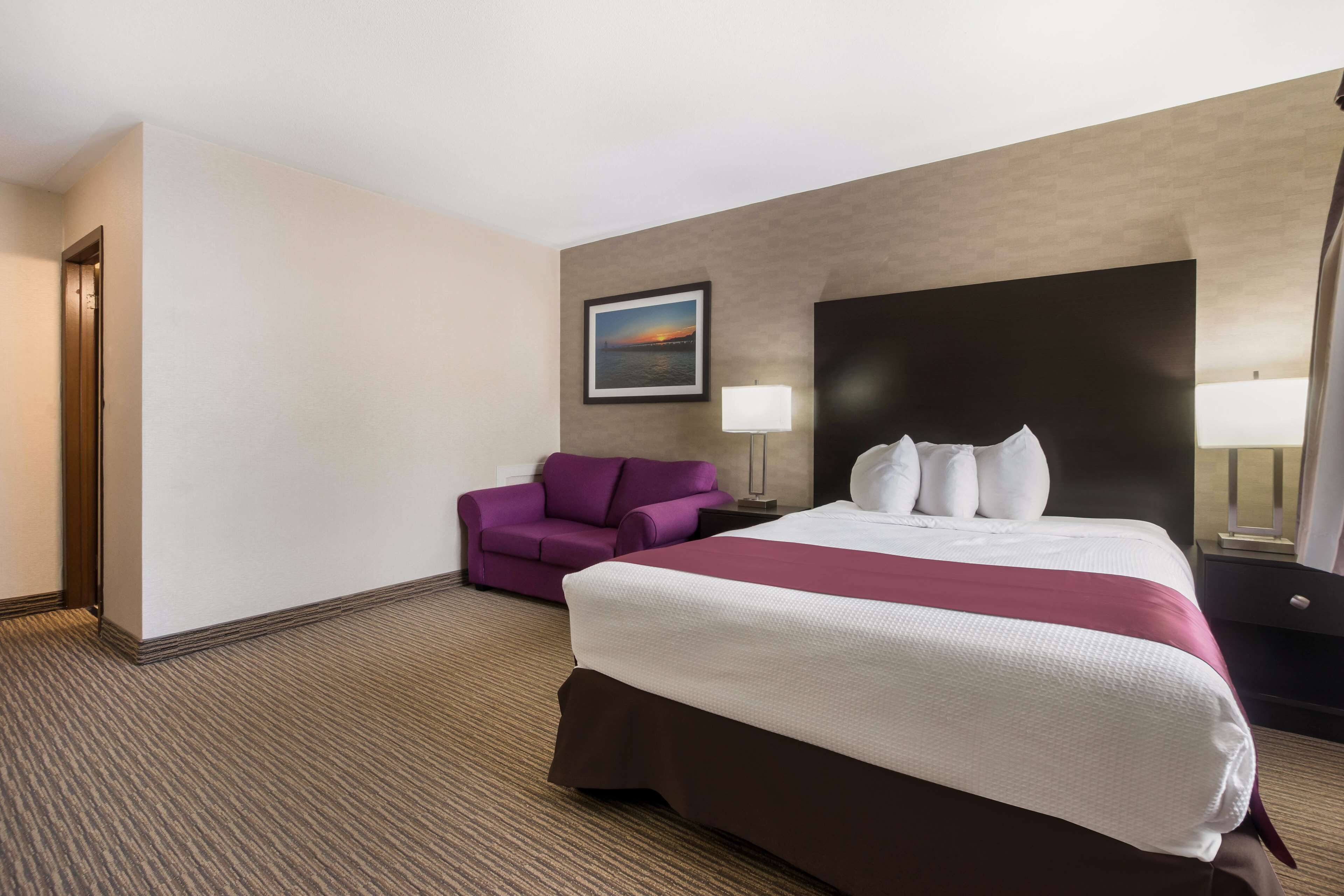 Una habitación de hotel con una cama grande, un sillón púrpura, una mesita de noche con lámpara y un cuadro en la pared.