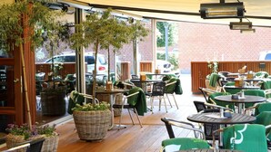 An outdoor seating area with wicker tables and chairs, potted plants, and a view of the street.