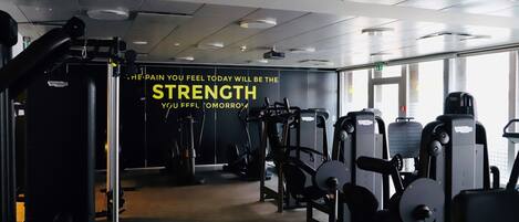 A gym with various exercise machines, a motivational quote on the wall, and large windows allowing natural light.