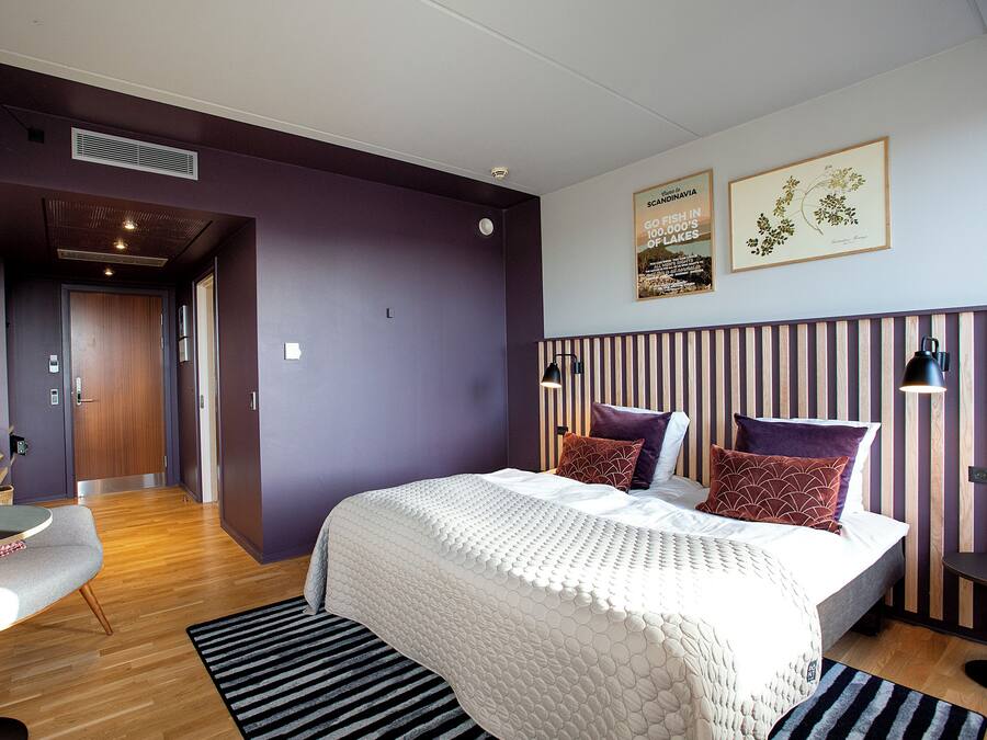 Habitación de hotel moderna con una cama grande, un escritorio y una silla. Hay dos cuadros en la pared y una ventana con persianas.