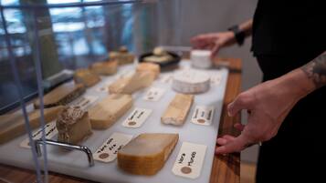 A display of various cheeses on a table with labels, a person reaching for one, and a glass display case in the background.